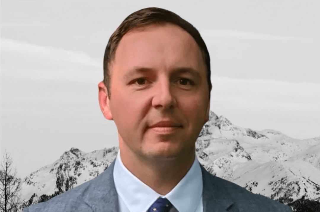 Portrait of Kevin Pavnica in a gray suit and tie, with snow-covered mountains behind him