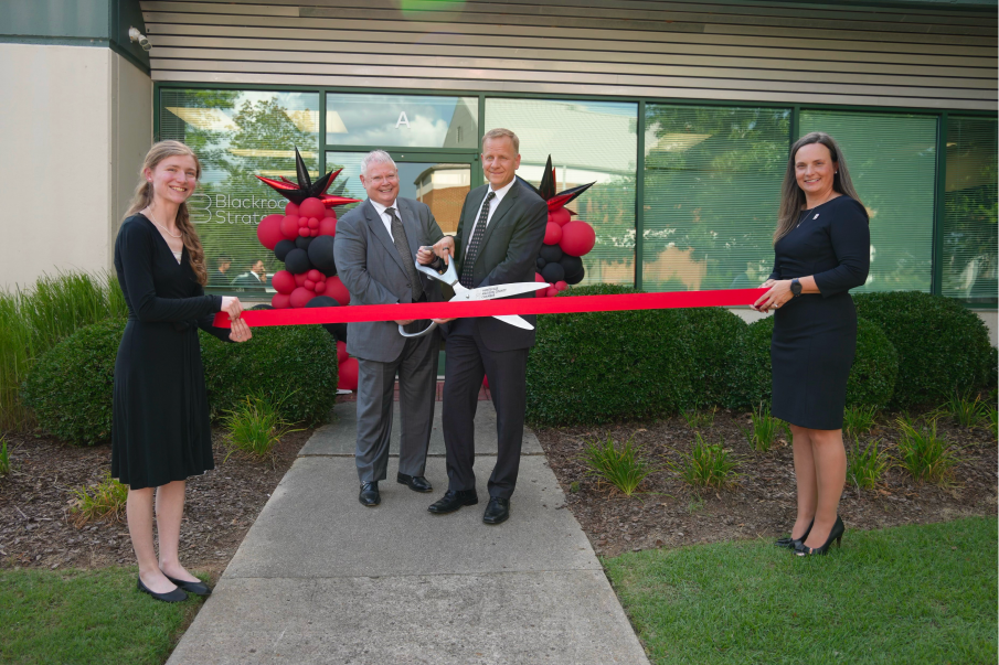 Two men cut a red ribbon with oversized scissors while two women hold its ends, outside an office entrance with red and black balloon arches