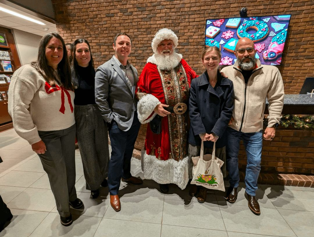 A group of people pose with Santa Claus in front of a brick wall and a screen showing decorated holiday cookies