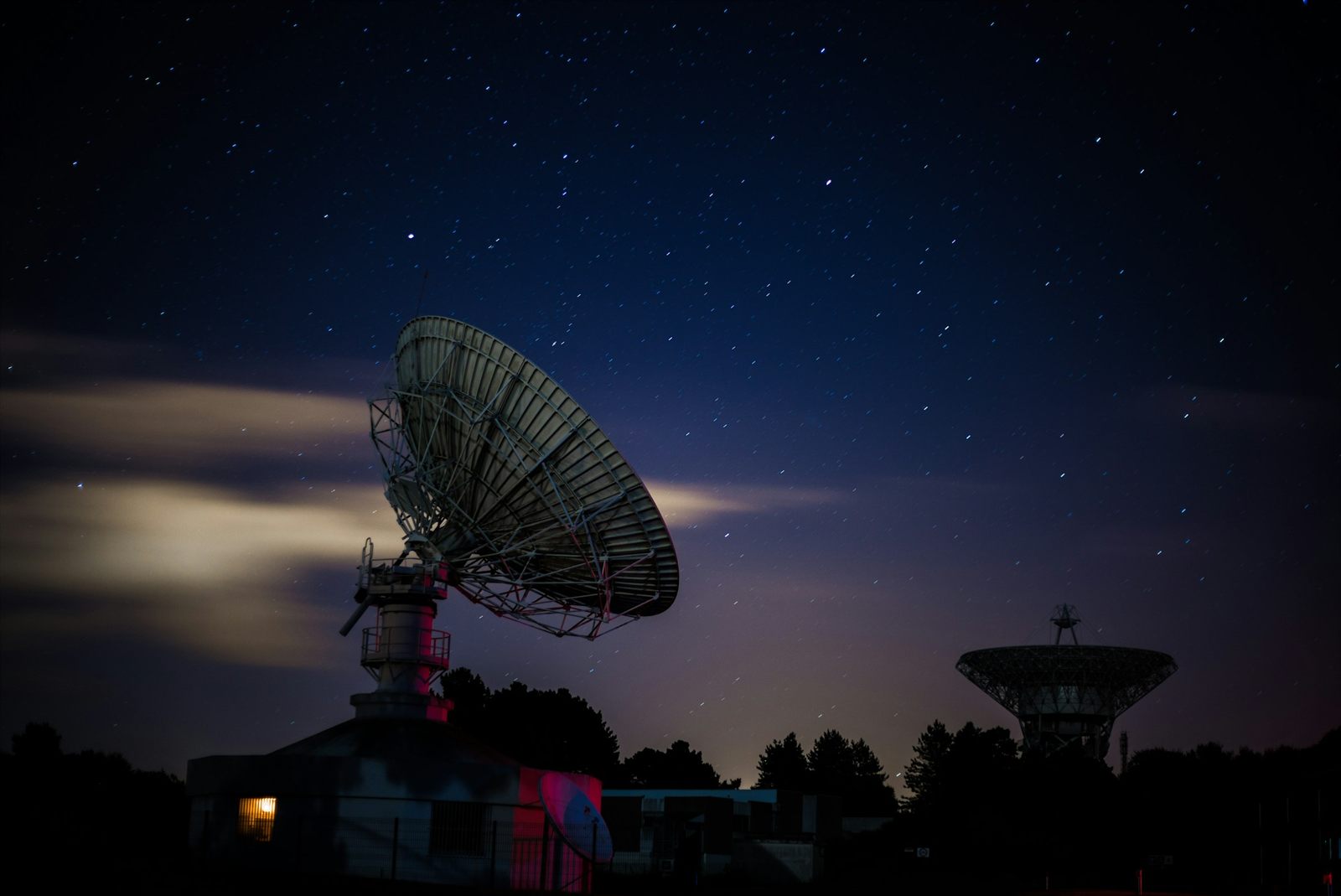 Two large dish antennas under a starry night sky, the nearer one beside a low building with a lit window