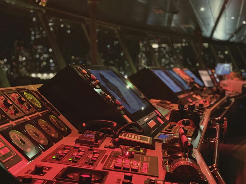 Red-lit consoles, gauges and screens on a ship's bridge at night, with city lights beyond the windows