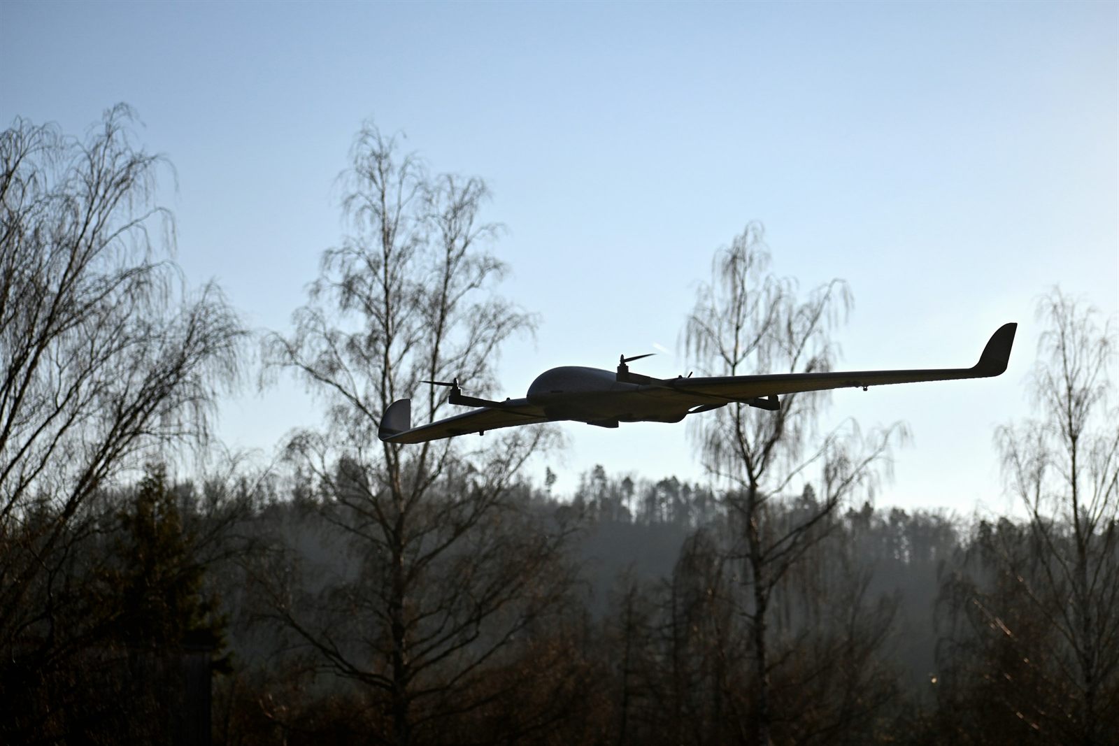 A fixed-wing drone with lift rotors on booms under its wings, flying past bare birch trees against a pale sky